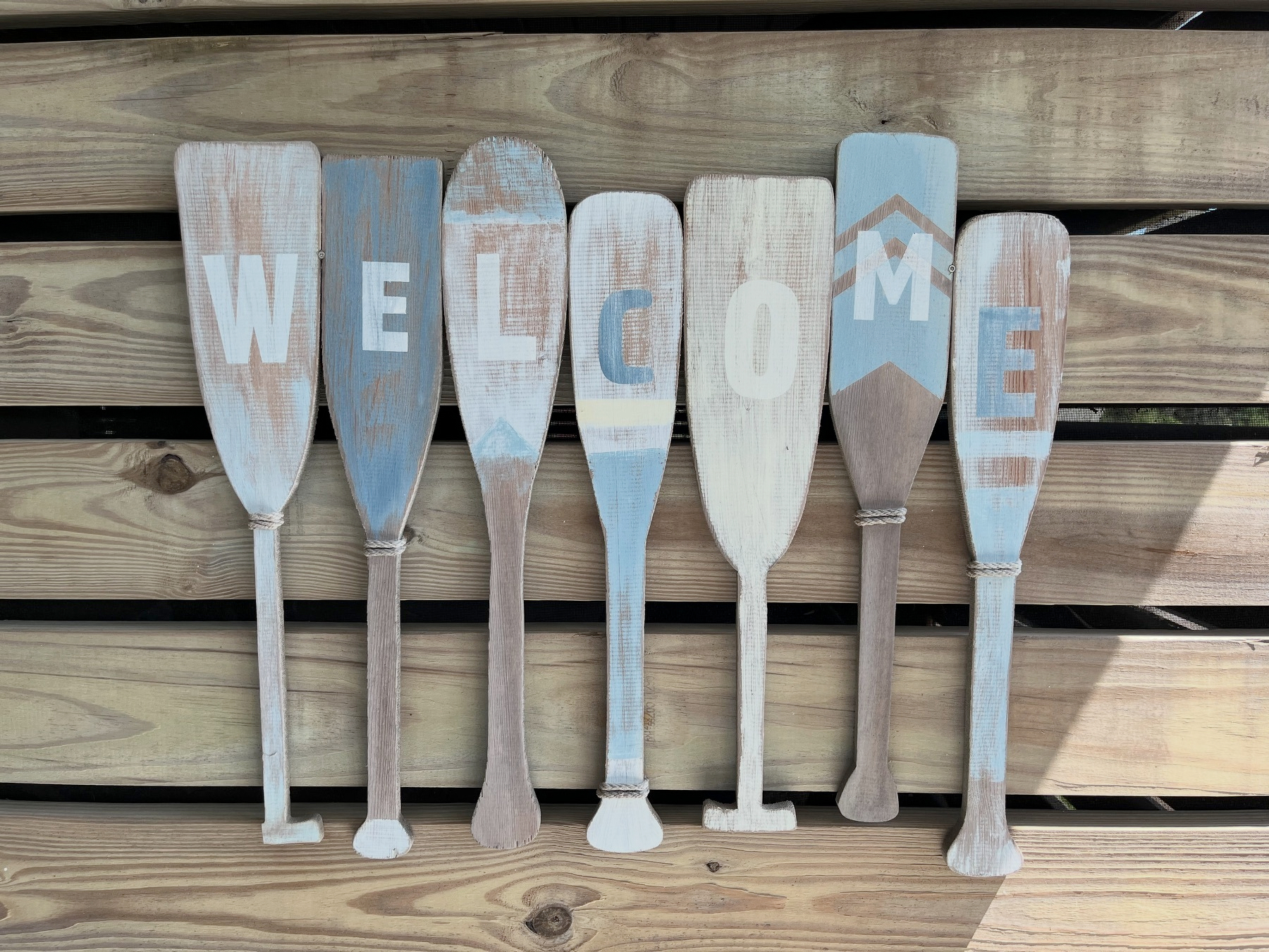 Seven wooden paddles painted in soft blue and white tones are arranged side by side to spell the word WELCOME.