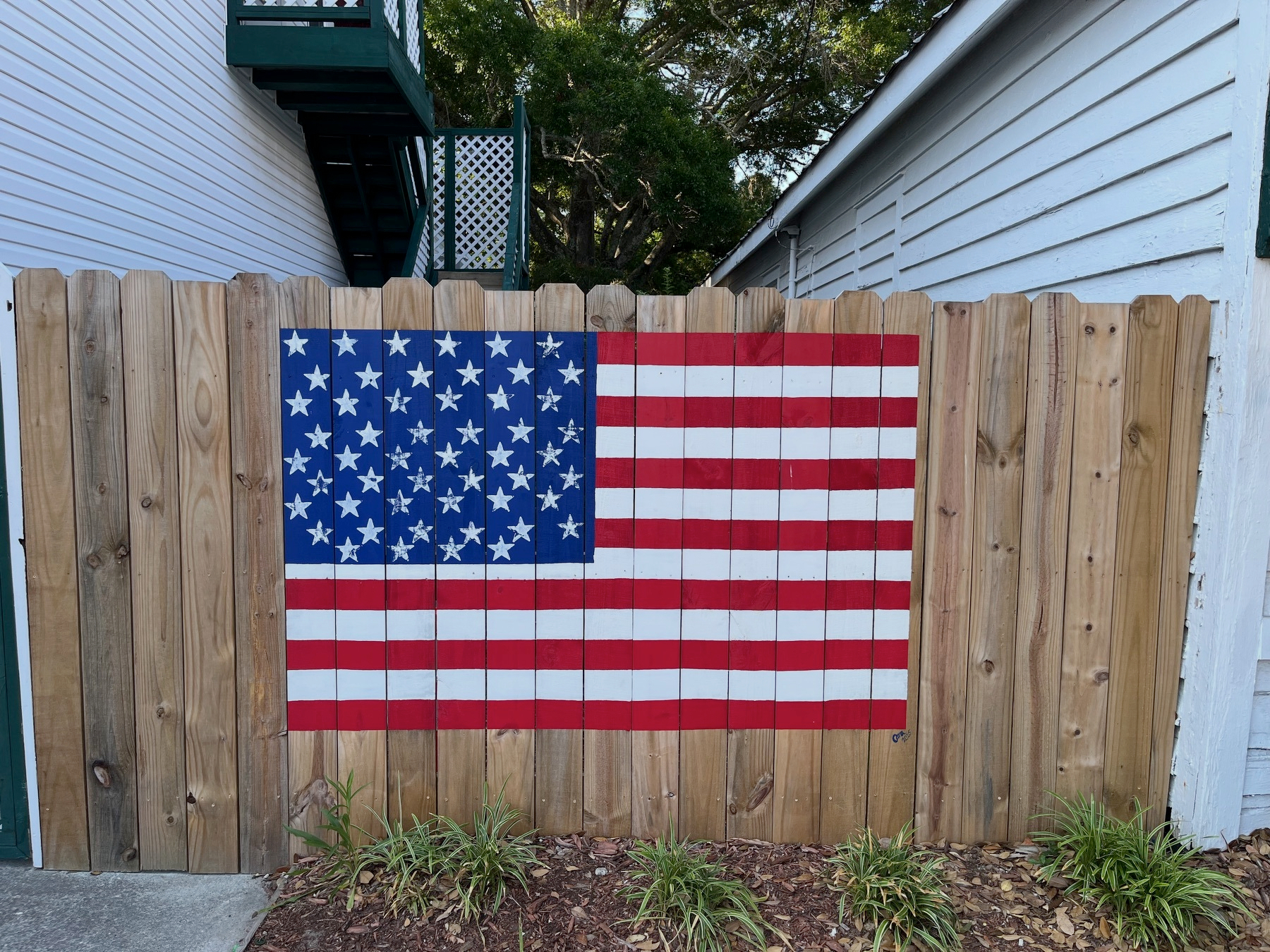 An American flag is painted on a wooden fence situated between two houses with some greenery at the base.