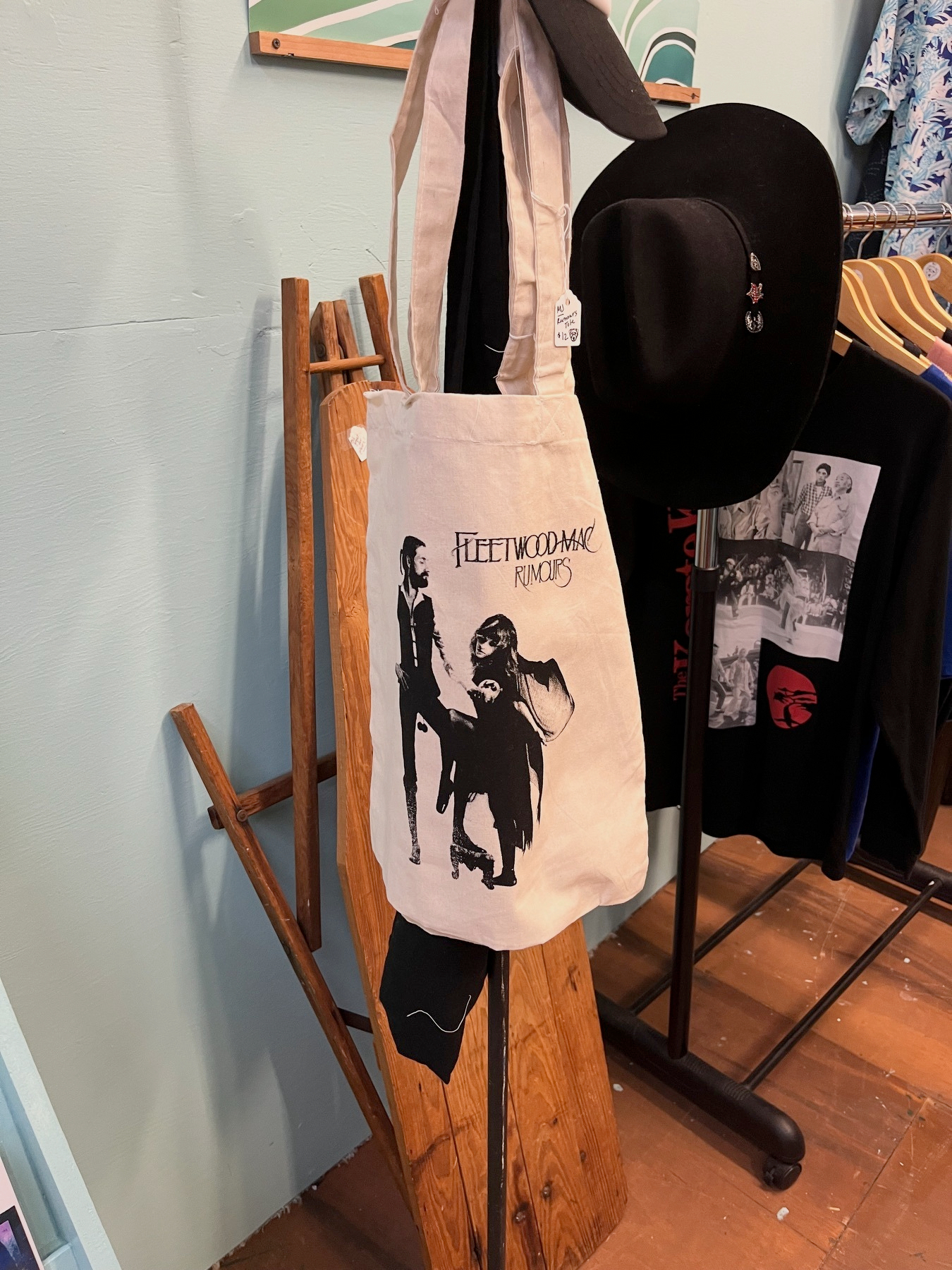 A beige tote bag featuring a Fleetwood Mac Rumors design is hanging on a wooden rack next to a black hat and some clothing.