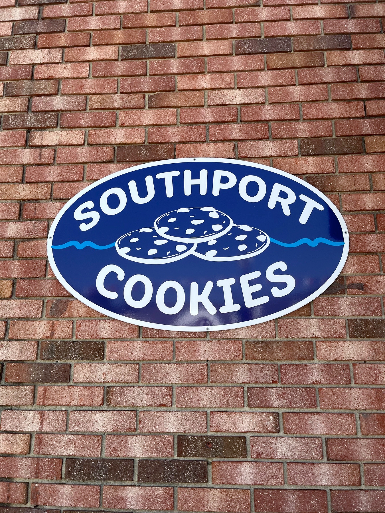 A blue oval sign with white text reading Southport Cookies and an illustration of three cookies is mounted on a red brick wall.