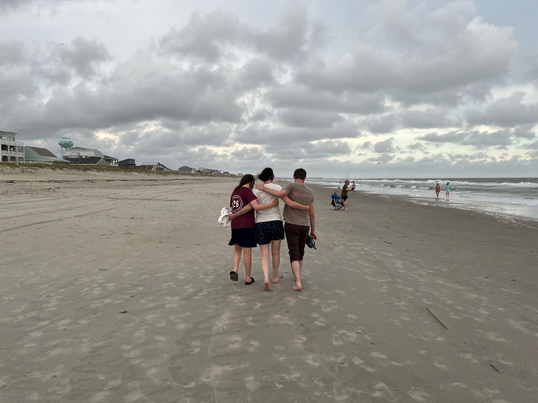 Three people walk arm in arm along a sandy beach under a cloudy sky.